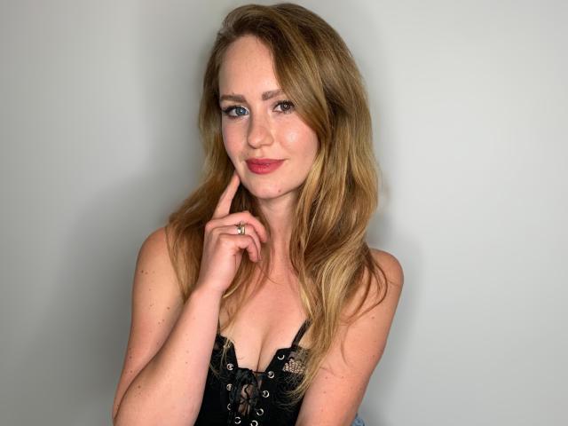 LyraFantasy with blonde hair, blue eyes, and a cute smile, touching her finger to her cheek, wearing a black lace-up top, showing her tits.