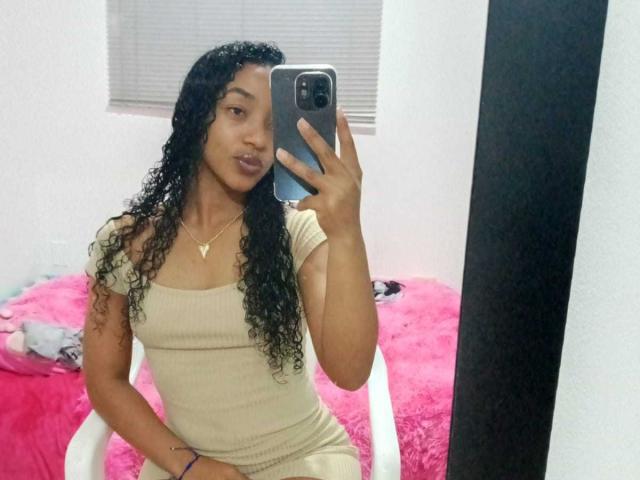 AlonnaGonzales with curly black hair, medium brown skin, wearing a beige ribbed dress, taking a mirror selfie with a gold necklace, showing her pouty lips, sitting on a pink chair.