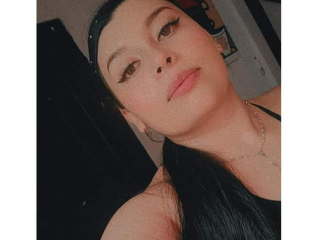 EmiliSimi has dark hair, brown eyes, and full lips. She's wearing a black top, hoop earrings, and a necklace. Her makeup includes thick, pointed eyeliner.