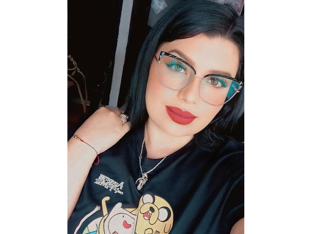 EmiliSimi, con cabello negro, sombra de ojos verde, lápiz labial rojo, gafas, una camisa negra con un dibujo animado, sosteniendo la camisa, un collar, un anillo y una pulsera, mirando a la cámara. Sus senos son ligeramente visibles..