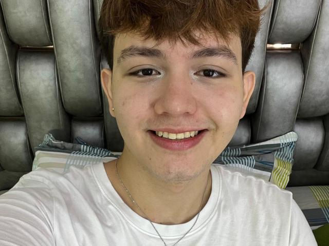 ErosDelRey has short brown hair, a slight smile, and is wearing a white tee with a silver necklace. He has small, barely noticeable tits and a cute face.