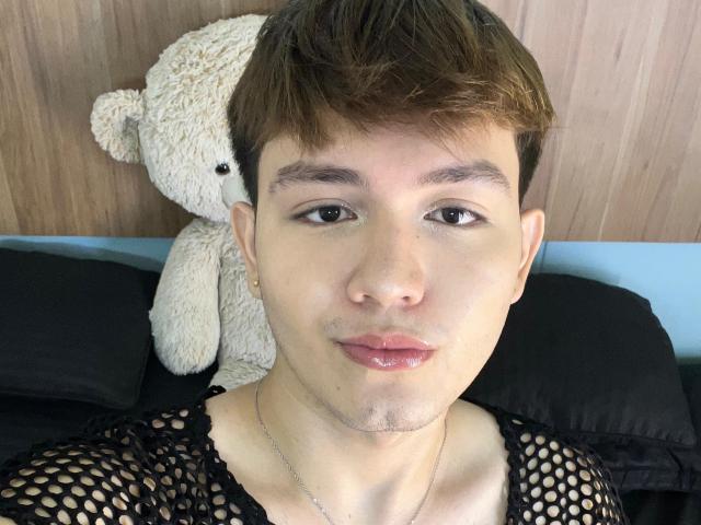 ErosDelRey has short brown hair, a light skin tone, and glossy pink lips. He's wearing a black fishnet shirt with a white stuffed teddy bear behind him.