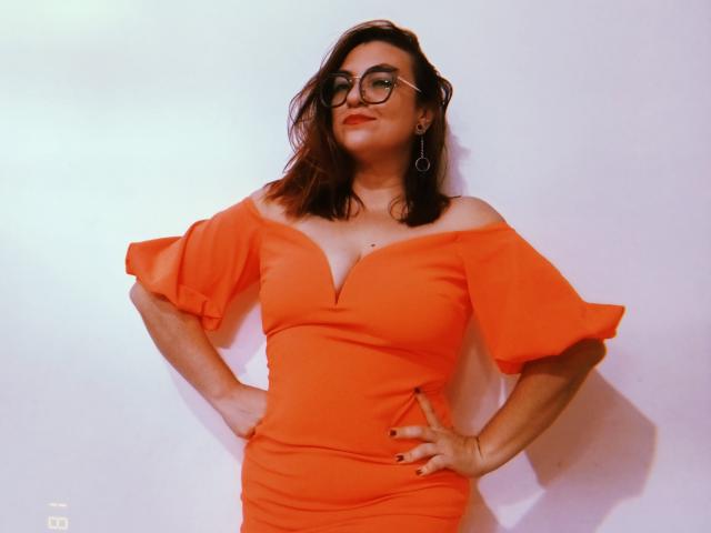 LunaRausch with red-orange dress, puffy sleeves, and cleavage, wearing glasses, earrings, hands on hips, slight smile, wavy brown hair, white wall, looking confident.