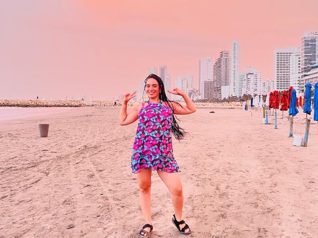 Hera, uma deusa com longos cabelos pretos, vestindo um vestido floral rosa e azul, em pé em uma praia de areia, fazendo gestos com as mãos no estilo rock and roll, sorrindo, usando sandálias pretas..