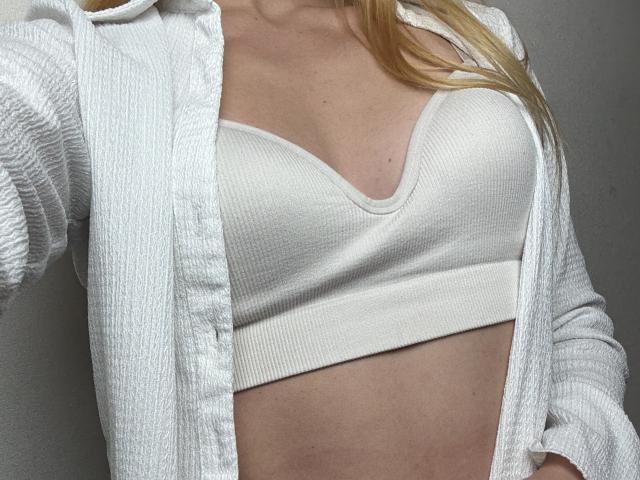 Twiksi vestindo um sutiã de renda branco, uma camisa branca de textura waffle aberta, cabelo loiro, pele clara, mostrando um pouco de sua barriga plana, tonificada e pálida. Os seios são de tamanho médio e visíveis..