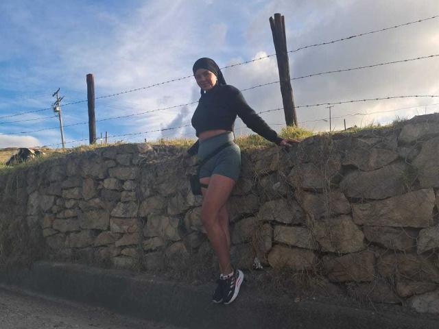 ArielleWilde with a black beanie, long-sleeve crop top, green shorts, and sneakers, leaning against a stone wall with barbed wire, and a clear blue sky.