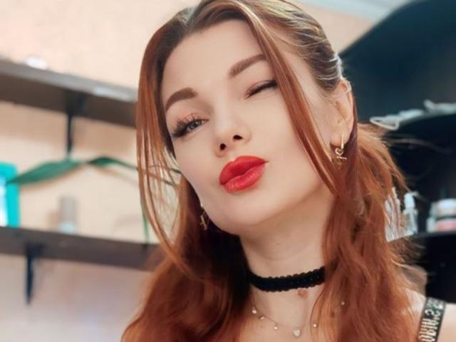 XRebeccaGlossX has red lipstick, winking, with long auburn hair, a black choker, and earrings, looking cute with slightly pouty lips. She's wearing a silver necklace.