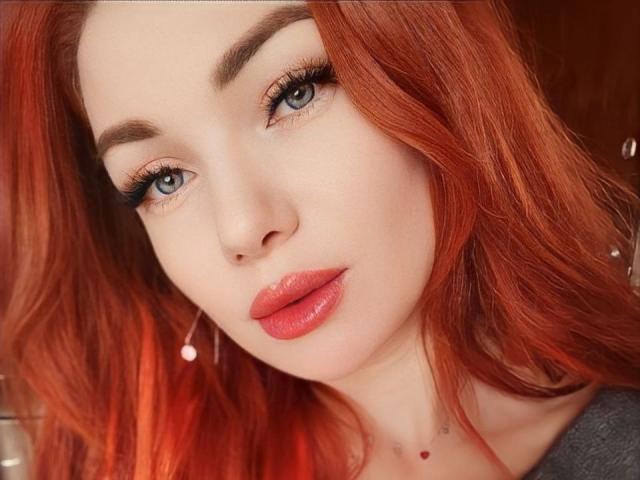 XRebeccaGlossX has red hair, blue eyes, full pink lips, and a cute face with subtle makeup. Her tits are not visible. She's wearing small, dangling earrings.