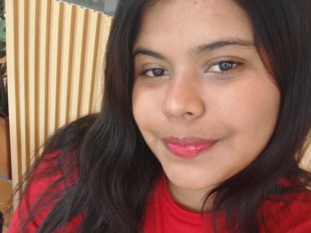 TatyM hat eine mittelbraune Haut, schwarzes, welliges Haar, braune Augen und roten Lippenstift. Sie trägt ein rotes Hemd, lächelt leicht, hat eine dezente Nase und kein sichtbares Make-up..