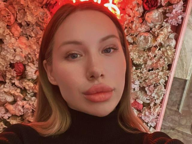 BellaLuxix has puffy pink lips, blue eyes, and blonde hair, wearing a black turtleneck, with a floral wall and red neon light in the background.