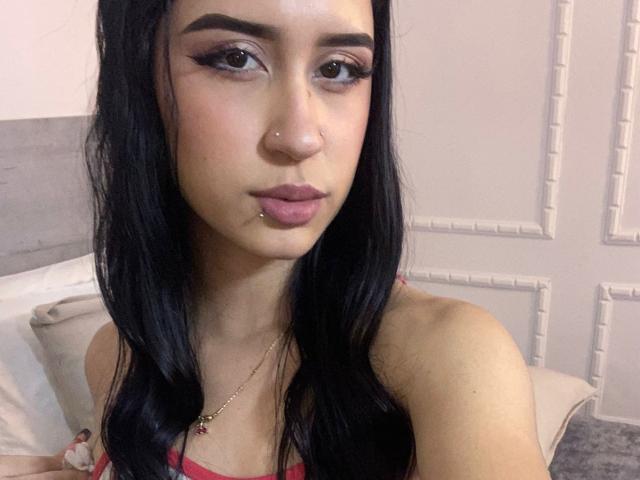 AnnaTorress, con cabello negro largo, nariz perforada y aro en el labio, vestida con una blusa roja, mirando a la cámara, mostrando sus senos, con maquillaje rosa suave, un collar dorado, y una cama en el fondo..