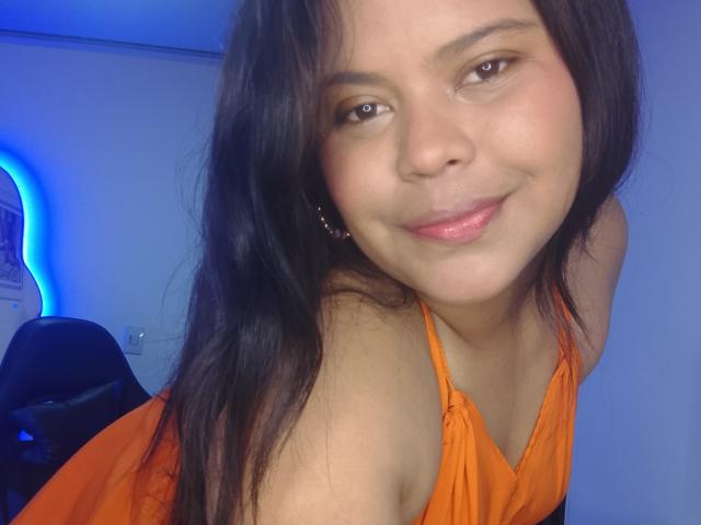 TatyM, con cabello negro largo, ojos marrones y una sonrisa encantadora, vestida con un top naranja de tirantes, inclinándose hacia adelante, mostrando sus pequeños senos y labios rosados. Luz azul en el fondo..