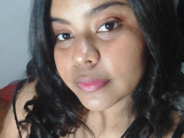 TatyM tiene ojos marrones, cabello negro rizado y labios rosados con un ligero brillo. Lleva una blusa negra y mira directamente a la cámara con una expresión neutra..