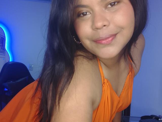 TatyM, avec ses longs cheveux noirs, sa robe orange et son rouge à lèvres rose, sourit doucement à la caméra, montrant légèrement ses seins et ses fesses. Une lumière bleue est visible en arrière-plan..