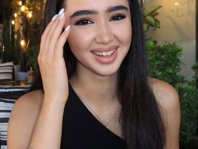 Lali69x with long black hair, smiling with pink lips, wearing a black one-shoulder top, touching her face with white-manicured nails, brown eyes, and a silver necklace.