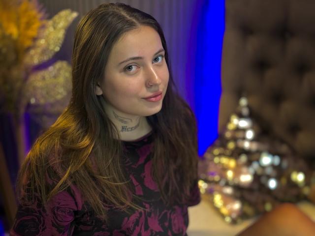 LilaVame, com cabelos castanhos longos, vestindo um vestido preto com estampas florais, sorrindo discretamente, olhos azuis, nariz pequeno, tatuagem no pescoço, sentada em frente a uma cabeceira marrom e felpuda, ligeiramente desfocada..