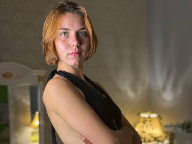 HannaDiya with short blonde hair, fair skin, small tits, and a black top, arms crossed, standing in a softly lit room, looking directly at the camera, serious expression.