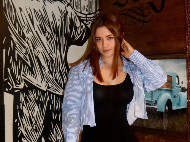 MaidieTrissy with brown hair, wearing a blue striped shirt over a black tank top, standing in front of a black and white sketch, hand in hair, looking at camera.