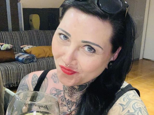 StrengeTante-hot has pale skin, black hair, blue eyes, red lipstick, tattoos, nose ring, black tank top, and holds a glass, with sunglasses on her head.