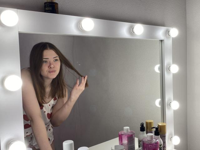CyberAlisa, avec des cheveux bruns, tenant une serrure, portant un débardeur blanc avec des cœurs rouges, debout devant un miroir avec des lumières rondes, entourée de produits de maquillage et de flacons..