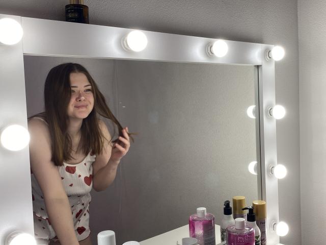 CyberAlisa with brown hair, wearing a white tank top with red heart patterns, stands in front of a mirror with round bulbs, touching her hair, surrounded by makeup.