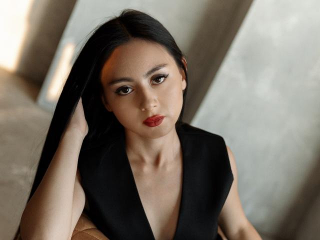 MiyaStiverts has long black hair, red lipstick, and a black sleeveless top, looking serious with her hand in her hair, showing her tits slightly in a soft light.