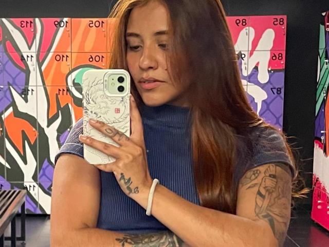 AlanaQuin with brown hair, blue ribbed turtleneck, tattoos on arm, holding white phone with floral case, taking selfie, looking down, pink lips, wrist bracelet.