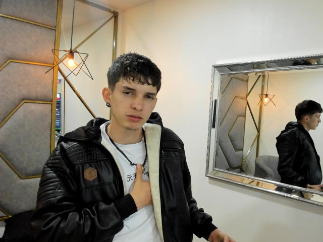JheamPiero with short black hair, black leather jacket, white shirt, and ear gauges, standing in front of a mirror, showing a neutral expression, with a geometric lamp behind.