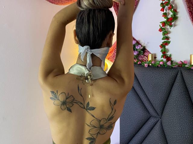 DalilaAliX with arms up, back to camera, silver hair in bun, white mask, floral tattoo, exposed back, grey fabric, floral garland on right.