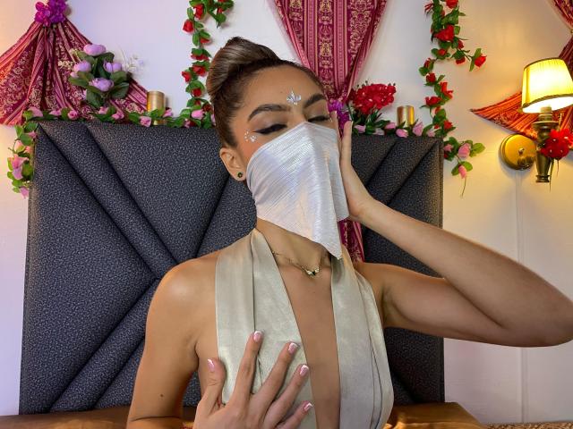 DalilaAliX with brown hair in a bun, wearing a silver mask, beige halter top, and glitter on her forehead, touching her mask with her right hand, showing her tits.