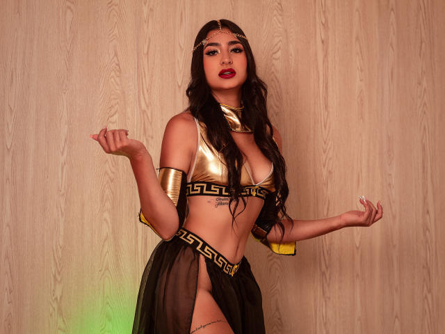 JasmineRh with long black hair, gold headpiece, gold bra, black and gold Grecian-style skirt, gold arm bands, and red lipstick, showing her tits and a tattoo on her belly.