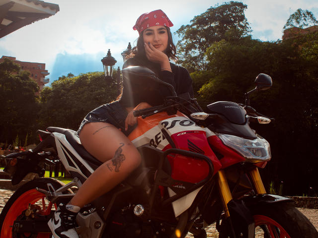 JasmineRh with red bandana, black top, and short black shorts, sitting on a red and white motorcycle, showing a tattoo on her right thigh, smiling, hands near face.