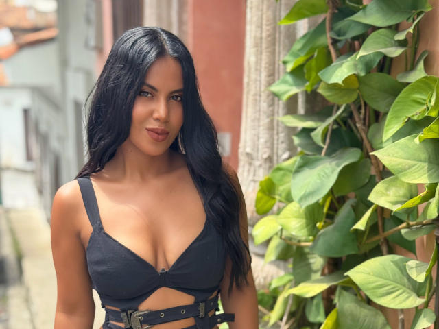CaraVelvet has black hair, tan skin, and wears a black bra with straps and a belt. She's standing next to green leaves, with a slight smile. Boobs are visible.
