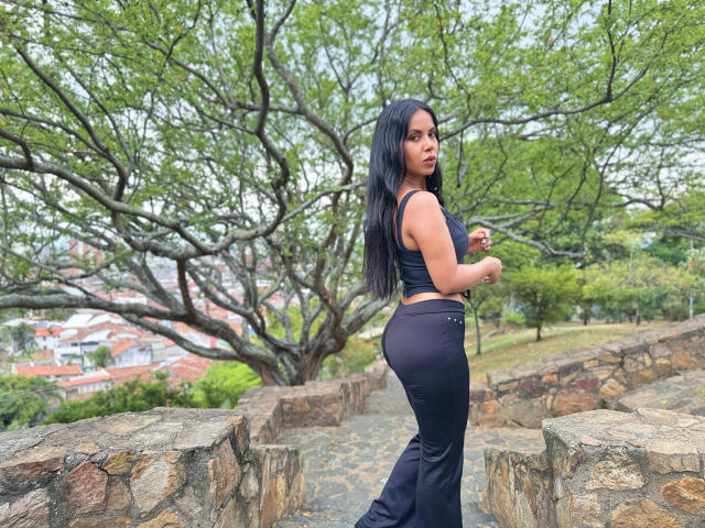 CaraVelvet has long black hair, wearing a black crop top and high-waisted black pants, showing her ass while looking over her shoulder on a stone pathway.