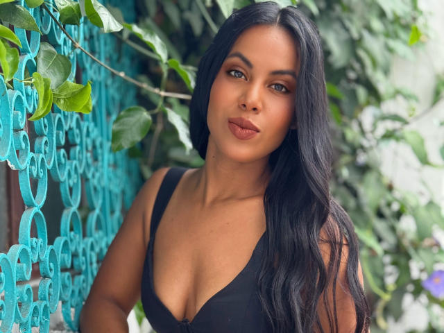 CaraVelvet with long black hair, brown eyes, and full lips, wearing a black bra, showing her tits, standing by a teal metal fence with green leaves.