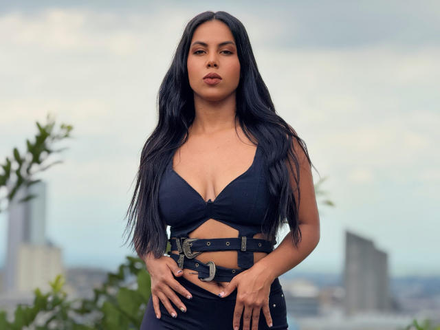 CaraVelvet has long black hair, medium tan skin, and wears a black bra with cutouts and a belt. Her hands rest on her hips, showing her tits.