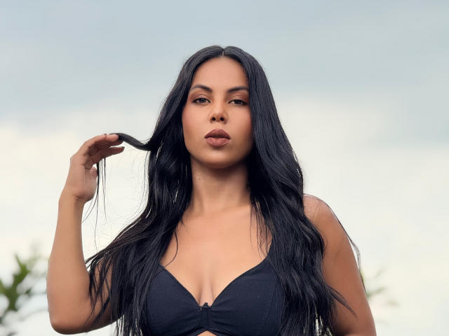 CaraVelvet has long black hair, a serious face, and is wearing a black bra. She's holding a lock of hair with her right hand, pouting slightly.