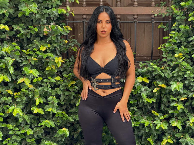 CaraVelvet with long black hair, wearing a black crop top with straps and black pants, standing in front of green leafy vines, showing her tits and a small ass.