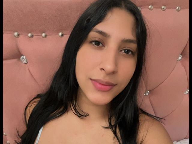 MadisonReyes has long black hair, brown eyes, and pink lips, with a cute smile. She's wearing a white top, and the pink, button-tufted headboard is in the background.