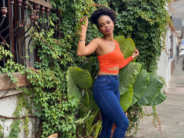 ChantalHaneke with curly black hair, orange one-shoulder top, and blue jeans, standing against a green leafy wall, hand in hair, looking down, showing her ass and small tits.