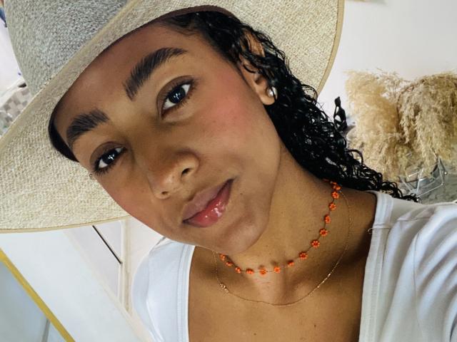NatalyBlonder has dark brown skin, curly black hair, and wears a straw hat, white top, and orange bead necklace. Her lips are slightly parted with a subtle smile.