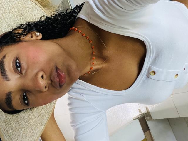 NatalyBlonder with dark curly hair, brown skin, and pink lips, wearing a white top with buttons and an orange bead necklace, showing small tits and a gold necklace.