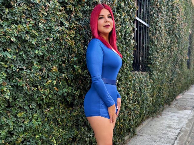 CorineRay with red hair, wearing a tight blue dress, showing her ass and tits, standing against a leafy wall, looking at the camera with a slight smile.