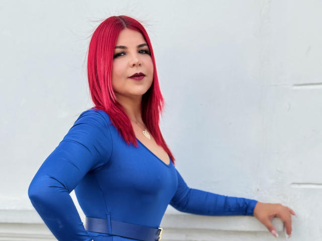 CorineRay with bright red hair, wearing a tight blue dress, showing her tits, standing with one hand on a white wall, looking confident with dark red lipstick.