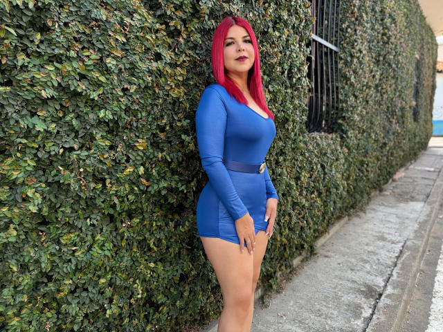 CorineRay with bright red hair, blue dress, and black belt, standing against a green hedge, showing her curvy ass and big tits. She's looking at the camera, smiling.
