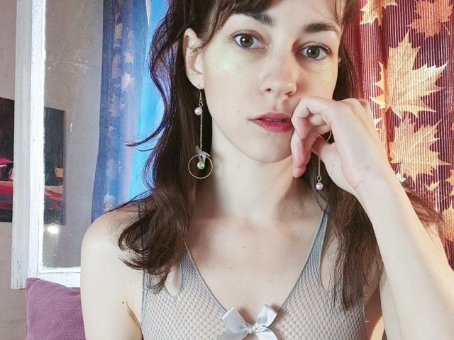 Barbariana with pale skin, brown hair, wearing a gray mesh top with a small white bow, green earrings, and resting her chin on her hand, looking slightly bored.
