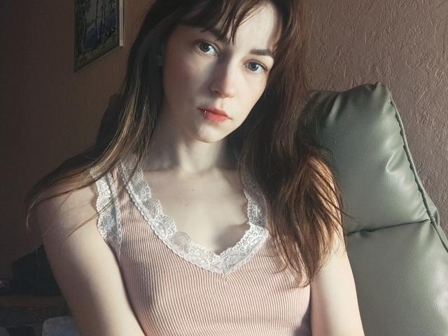 Barbariana with pale skin and brown hair, wearing a pink ribbed tank top with white lace trim, sitting in a green leather chair, looking slightly to the left.