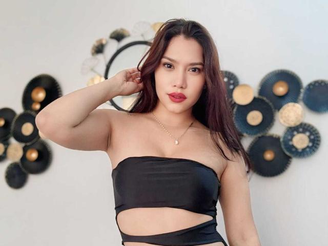 SoffieEssie with dark brown hair, wearing a black strapless top, showing her tits, with a hand in her hair, red lipstick, and a gold necklace.
