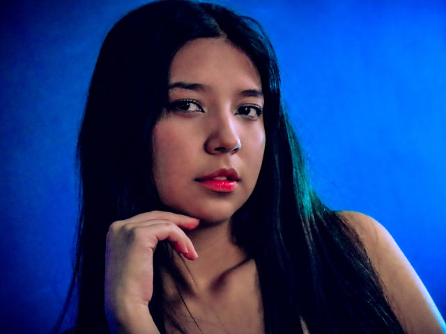 LayraNoir heeft lang zwart haar, rode lippenstift en een serieuze uitdrukking, met haar hand in de buurt van haar kin, tegen een blauwe achtergrond. Haar borsten zijn klein en haar achterkant is niet te zien..