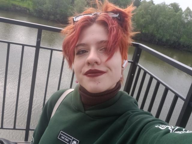LadyIlona with red messy hair, green sweater, and dark lipstick, standing by a river, smiling slightly, wearing glasses on head, shoulder bag visible, pale skin, small facial blemishes.